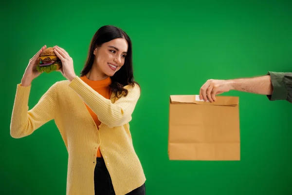 Чоловік тримає харчовий пакет біля усміхненої жінки зі смачним бургер ізольовано на зеленому — стокове фото