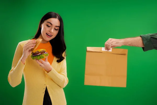 Задоволена жінка дивиться на смачний бургер біля доставки чоловік з паперовим мішком ізольовано на зеленому — стокове фото
