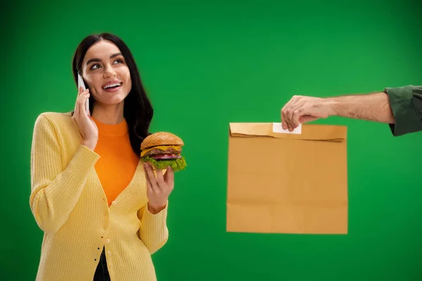 Усміхнена жінка тримає бургер і розмовляє на смартфоні біля кур'єра з паперовим мішком ізольовано на зеленому — стокове фото