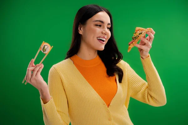 Pleased woman holding sushi roll and looking at tasty pizza isolated on green - foto de stock