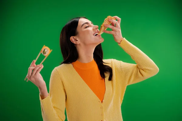 Young brunette woman holding chopsticks with sushi roll while eating pizza isolated on green — Photo de stock