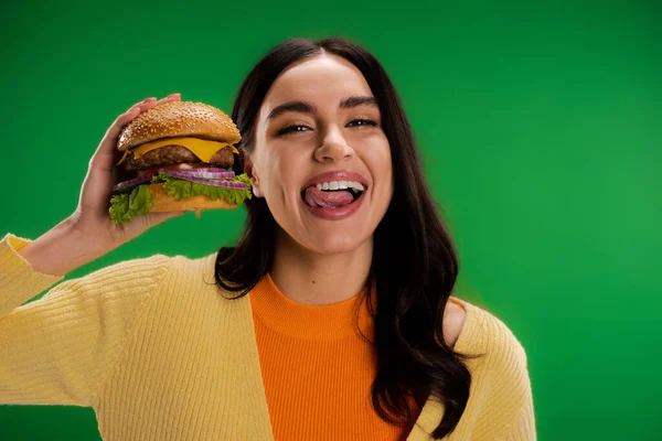 Brunette woman holding tasty burger and sticking out tongue isolated on green — Stock Photo