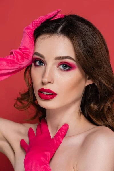 Portrait of brunette woman with magenta color eye makeup looking at camera while posing in gloves isolated on pink — Stock Photo