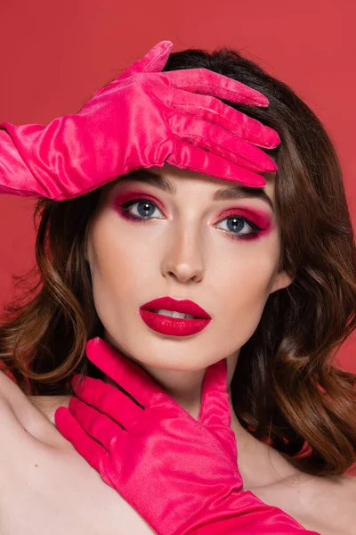 Young brunette woman with magenta color eye makeup looking at camera while posing in gloves isolated on pink - foto de stock