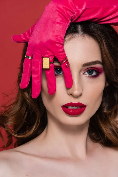 Brunette woman in magenta color glove with golden rings covering eye while looking at camera isolated on pink - foto de stock