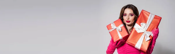 Brunette young woman in magenta color dress and gloves holding presents isolated on grey, banner — Stockfoto