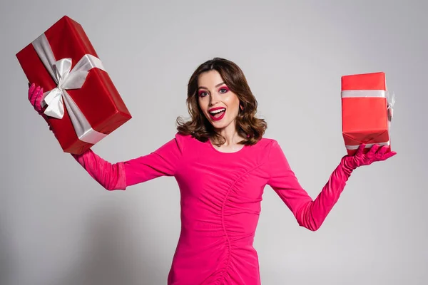 Amazed young woman in magenta color dress and gloves holding presents on grey — стоковое фото