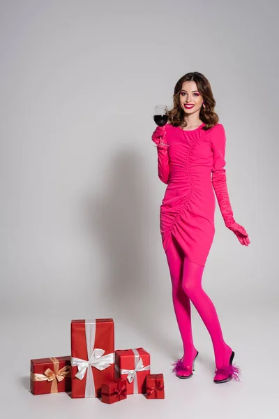 Full length of cheerful woman in magenta color dress holding glass with red wine near presents on grey — Foto stock
