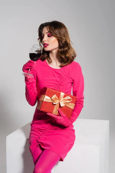 Young woman in magenta color dress sitting on white cube with glass of red wine and present isolated on grey — Photo de stock