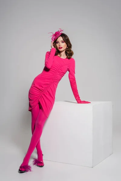 Full length of young woman in magenta color dress and elegant hat with feather posing near white cube on grey — стоковое фото