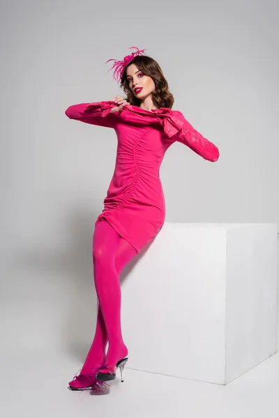 Full length of young woman in magenta color dress and hat with feather holding gloves near white cube on grey — Stock Photo