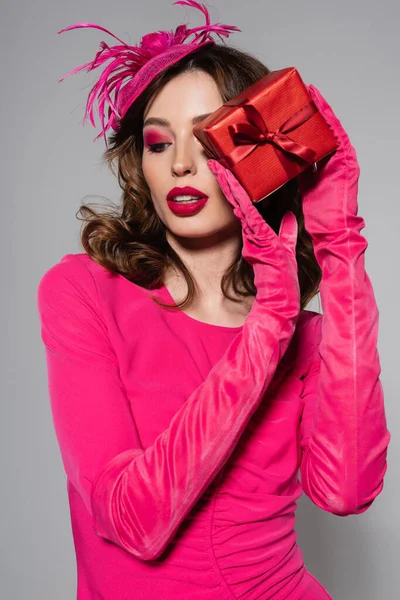 Brunette young woman in magenta color gloves and hat with feather holding gift box near face isolated on grey — Stock Photo