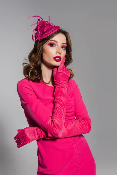 Pretty young woman in magenta color dress and hat with feather looking at camera isolated on grey — Fotografia de Stock