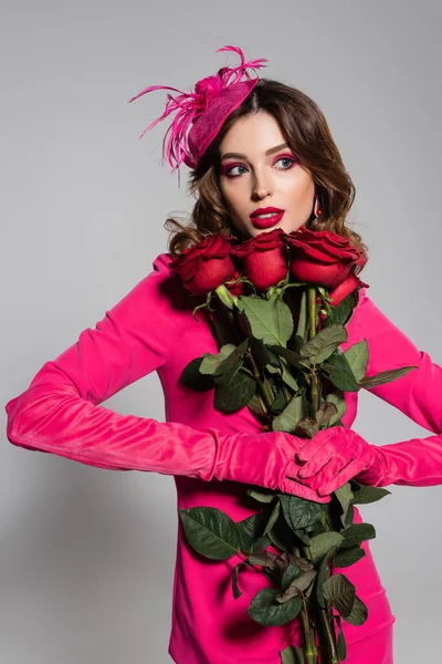 Brunette young woman in magenta color gloves and hat with feather holding red roses on grey — Photo de stock