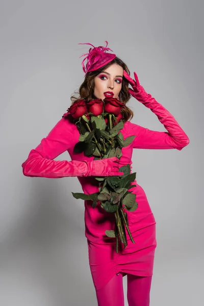 Brunette young woman in magenta color gloves adjusting hat with feather while holding red roses on grey — Stockfoto