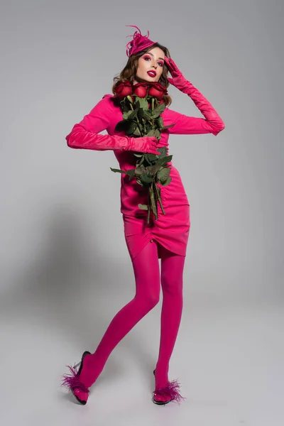Full length of young woman in magenta color gloves and hat with feather holding red roses on grey — Stock Photo