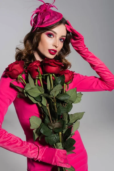 Young woman in magenta color gloves and hat with feather holding red roses isolated on grey — Foto stock