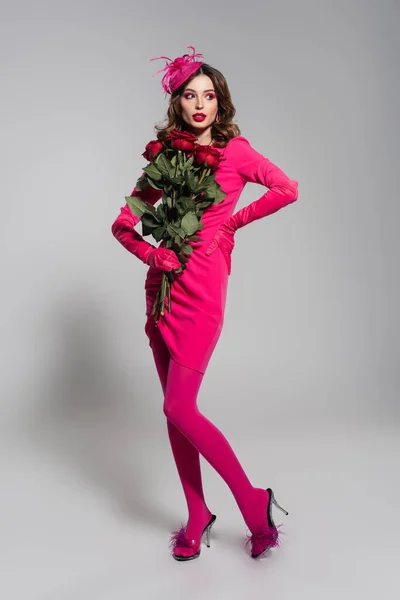Повна довжина молодої жінки в рукавичках кольору пурпурного кольору і капелюсі позує рукою на стегні, тримаючи червоні троянди на сірому — стокове фото
