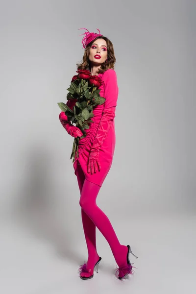Full length of young woman in magenta color gloves and elegant hat posing with red roses on grey — Stock Photo