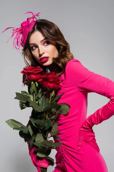 Brunette young woman in magenta color gloves and hat posing with hand on hip while holding red roses isolated on grey - foto de stock