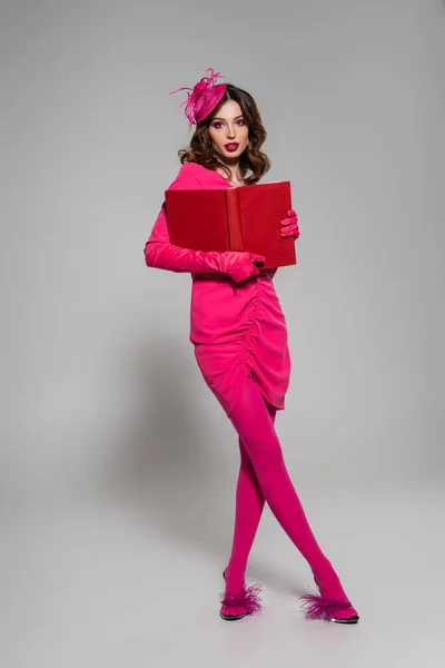 Full length of brunette woman in magenta color dress and hat with feather holding book on grey — Photo de stock
