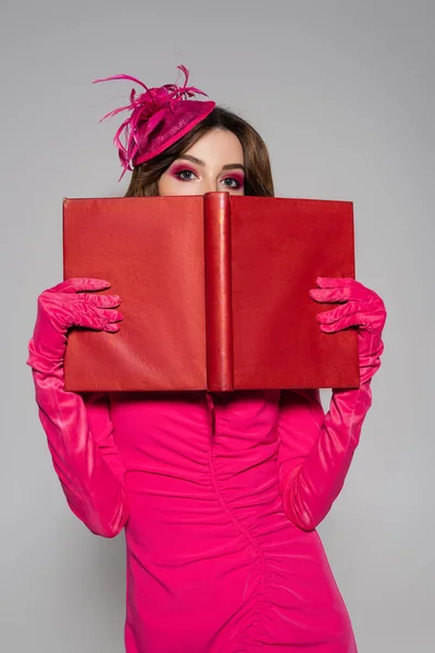 Young and brunette woman in magenta color gloves and hat with feather covering face with book isolated on grey — Stock Photo