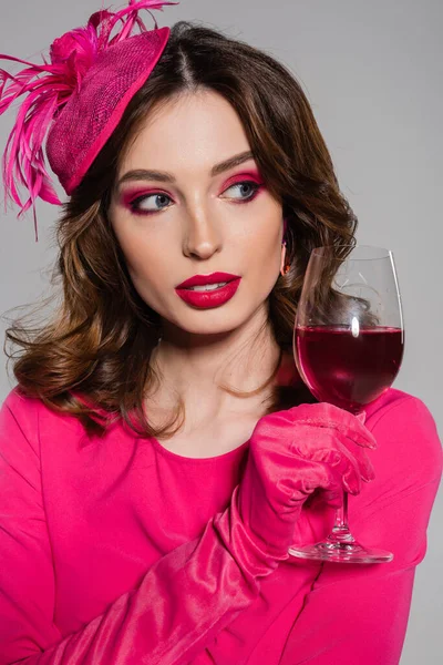 Portrait of young woman in elegant hat and magenta color dress holding glass with alcohol drink isolated on grey — Photo de stock