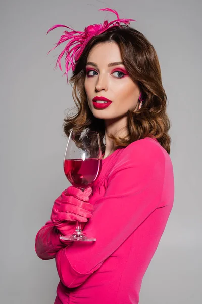 Portrait of brunette woman in elegant hat and magenta color dress holding glass with alcohol drink isolated on grey — Photo de stock