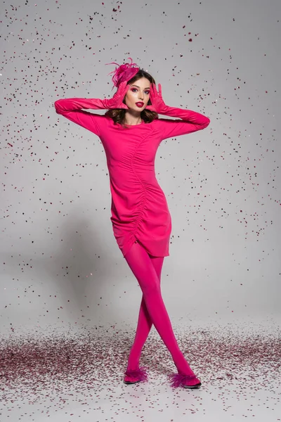 Full length of brunette woman in elegant hat and magenta color dress posing on grey with falling confetti - foto de stock