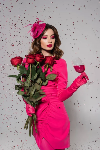 Brunette woman in elegant hat and magenta color dress holding roses and glass of wine on grey with falling confetti — Stock Photo