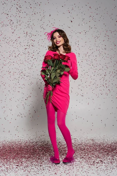 Full length of happy woman in hat and magenta color dress holding red roses on grey with falling confetti — Foto stock