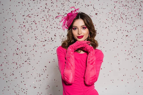 Cheerful woman in elegant hat and magenta color dress posing on grey background with falling confetti — Stock Photo