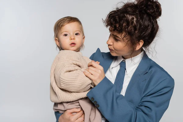 Curly businesswoman in suit with tie looking at baby daughter isolated on grey — Fotografia de Stock