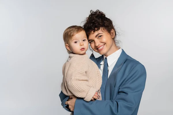 Femme d'affaires heureuse en costume avec cravate tenant dans les bras bébé fille isolé sur gris — Photo de stock