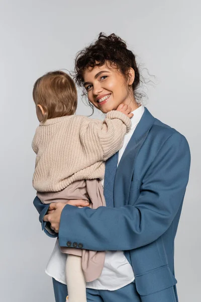 Cheerful businesswoman in blue suit holding in arms baby girl isolated on grey — Fotografia de Stock