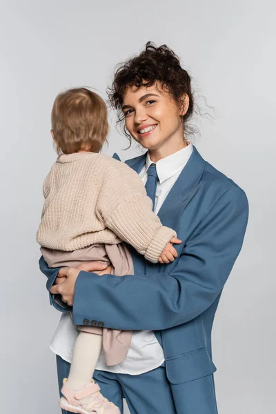 Happy businesswoman in blue suit holding in arms toddler daughter isolated on grey — Fotografia de Stock