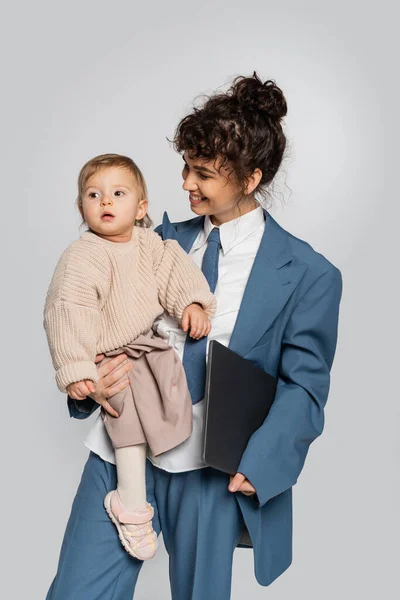 Cheerful businesswoman in blue suit holding in arms toddler daughter and laptop isolated on grey - foto de stock