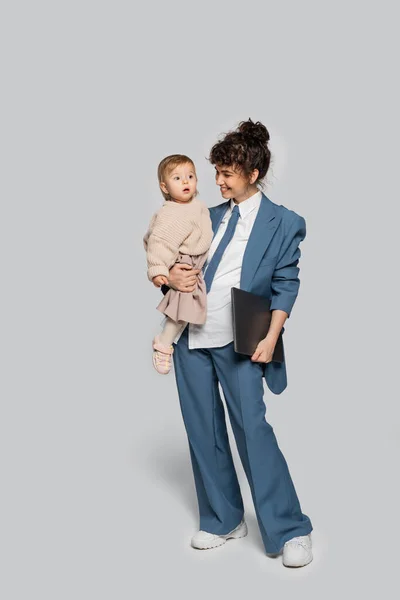 Full length of cheerful businesswoman in blue suit holding in arms toddler daughter and laptop on grey — Photo de stock