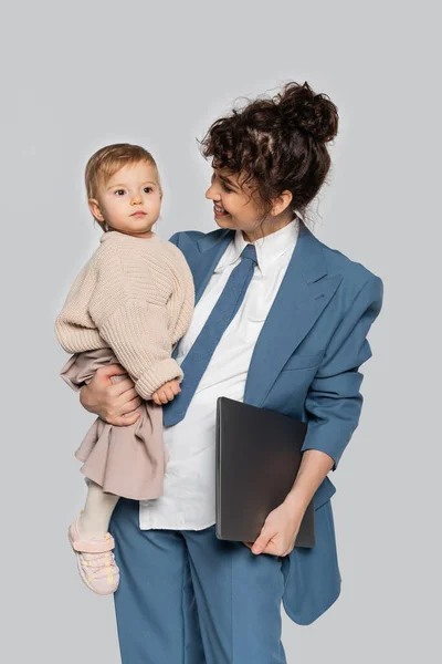 Happy businesswoman in blue formal wear holding in arms toddler daughter and laptop isolated on grey — Fotografia de Stock