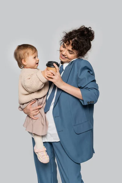 Happy businesswoman in blue formal wear talking on smartphone while holding toddler daughter and coffee to go isolated on grey — Stock Photo