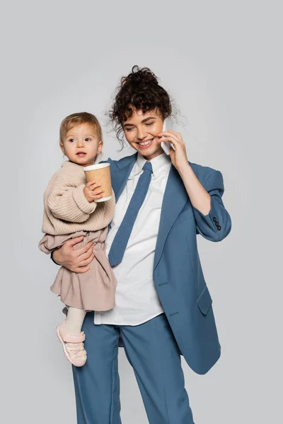 Happy businesswoman in suit talking on smartphone while holding toddler daughter with takeaway drink isolated on grey — Fotografia de Stock