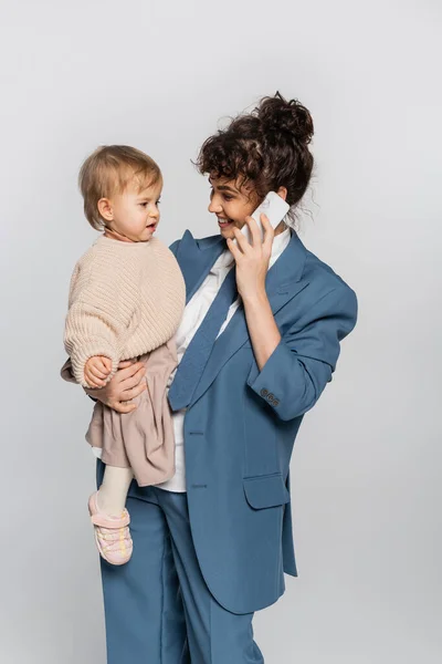 Happy businesswoman in suit talking on cellphone while holding toddler girl isolated on grey — Stockfoto