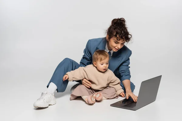 Curly businesswoman in suit sitting with toddler daughter and using laptop on grey — стоковое фото