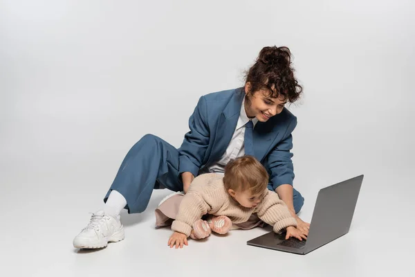 Happy businesswoman in suit sitting with toddler daughter and using laptop on grey — Fotografia de Stock