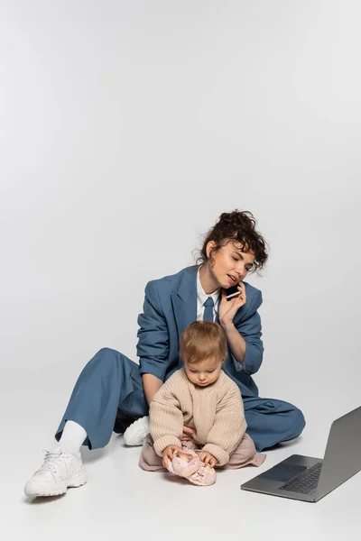 Busy businesswoman in suit talking on smartphone while sitting with toddler daughter near laptop on grey — Photo de stock