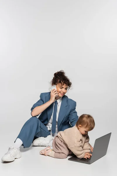 Femme d'affaires gaie en costume parlant sur smartphone tout en étant assis avec fille tout-petit près d'un ordinateur portable sur gris — Photo de stock