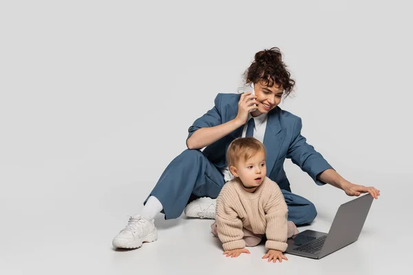 Happy businesswoman in suit talking on smartphone while sitting with toddler daughter near laptop on grey — Photo de stock