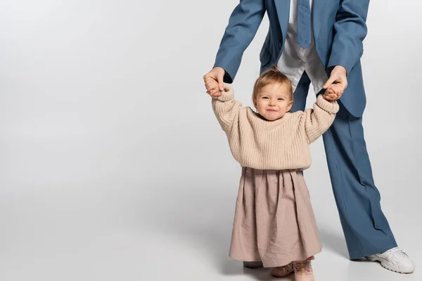 Mother in blue suit standing and holding hands of happy toddler girl on grey — Stock Photo