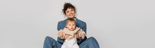 Happy businesswoman in blue suit sitting with toddler daughter isolated on grey, banner — Photo de stock