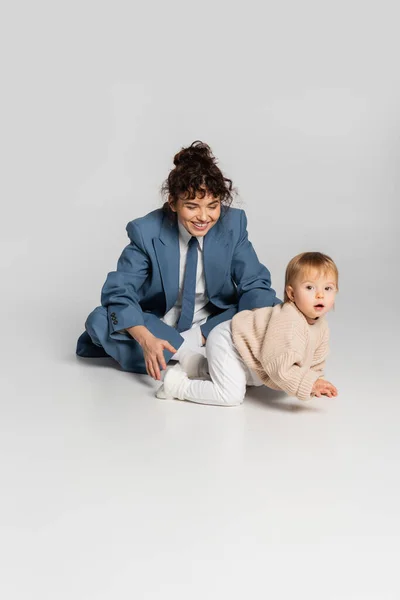 Curly businesswoman in blue suit looking at toddler daughter crawling on grey background - foto de stock